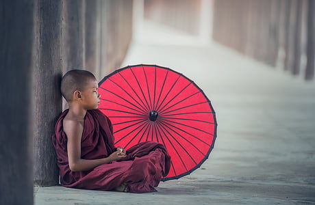 photo of child monk in maroon robe sitting on floor beside red parasol thumbnail