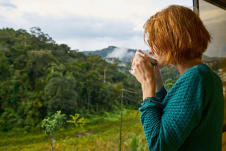 woman in blue long-sleeved top drinking coffee standing in front of mountains thumbnail