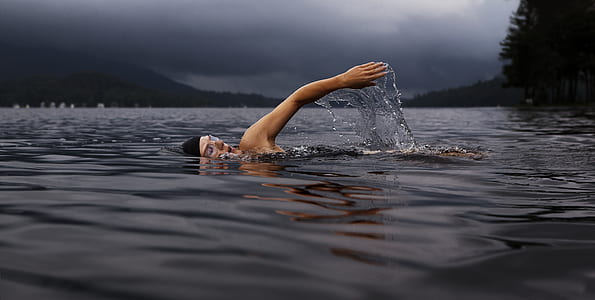 person swimming on body of water surrounded with tall trees under cloudy sky thumbnail