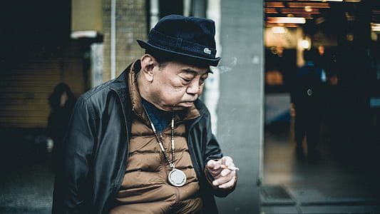 focus photography of man wearing black leather zip-up jacket holding cigarette while standing near building thumbnail