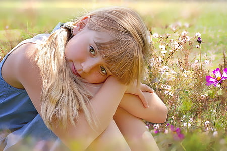 selective focus of girl wearing blue dress sitting on ground surrounded by flowers thumbnail