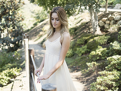 selective focus photography of woman in white dress holding on brown wooden fence while looking down thumbnail