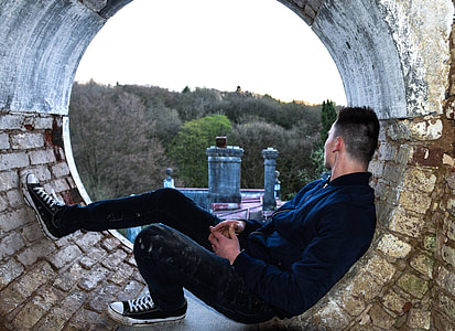 man wearing black jacket and black pants sitting on brown concrete surface with tall trees background thumbnail