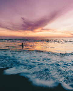 landscape photography of person standing on seashore surrounded by body of water during sunset thumbnail