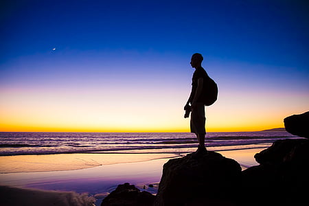 silhouette of photographer standing on rock looking at seashore during sunset thumbnail