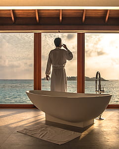 man wearing white bath robe standing next to free-standing bathtub inside bathroom near window during sunrise thumbnail