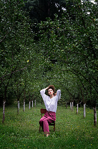 photo of woman in white long-sleeve top with red pants sitting on brown chair surround trees thumbnail