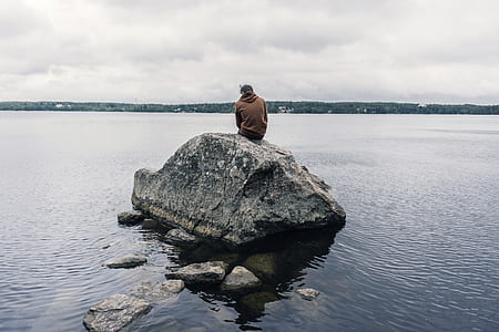 man sitting on large rock facing body of water thumbnail