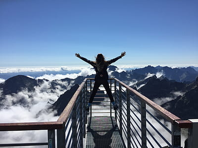 Person Standing on Hand Rails With Arms Wide Open Facing the Mountains and Clouds thumbnail