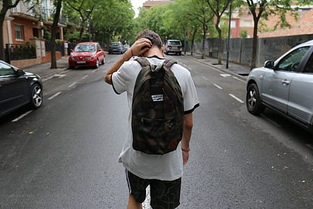 man wearing brown backpack walking on street between cars during daytime thumbnail