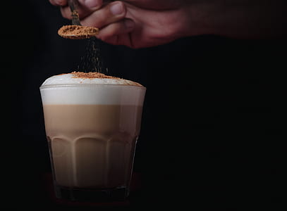 person's right hand holding spoon and slowly pouring brown powder on brown liquid filled clear drinking glass with bubbles on top thumbnail