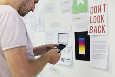 man using black smartphone while standing near white wall thumbnail