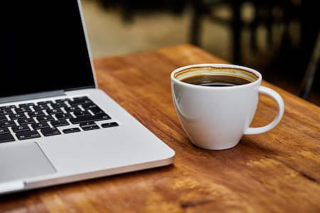 coffee filled white ceramic mug beside MacBook Pro on top of brown wooden table thumbnail