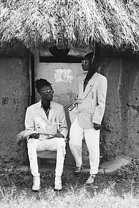 Grayscale Photo of Two Men Standing Near Hut thumbnail