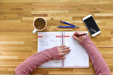 person writing on white notebook near white smartphone and cup on table thumbnail