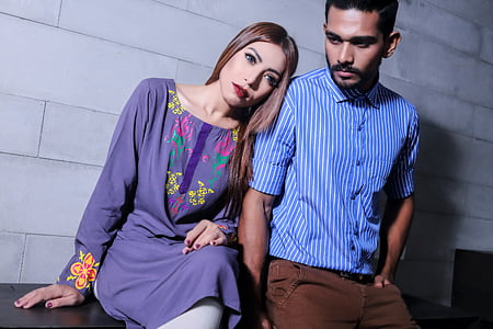 man in blue pinstriped dress shirt standing beside of woman in purple floral dress sitting on black wooden table and in front of gray wall thumbnail