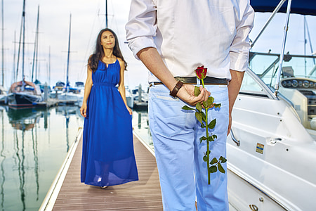 woman in blue sleeveless dress and man in white dress shirt and blue jeans holding red rose behind his back on pier bridge during daytime thumbnail