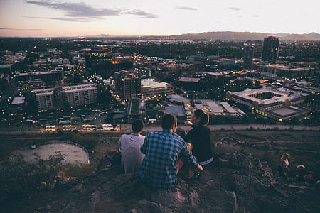 two men and one woman sitting on hill overlooking high rise buildings during daytime thumbnail