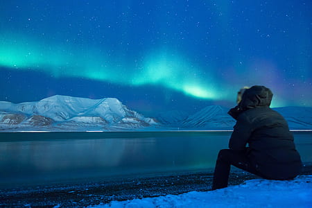person wearing black ski jacket sitting in front of body of water with distance at snowy hills under dark sky with Aurora borealis thumbnail