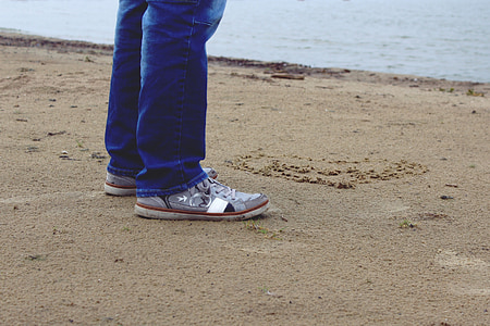 person in jeans and grey sneakers standing on beach sand thumbnail