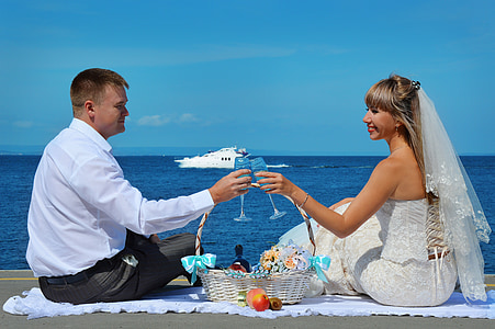 wedding couple holding wine glasses sitting beside seashore under blue sky during daytime thumbnail