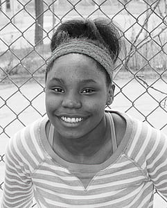 smiling woman wearing crew-neck top behind gray metal chain link fence thumbnail
