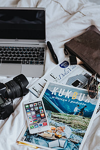 Silver laptop, a camera, magazines and other items on white bed sheets thumbnail