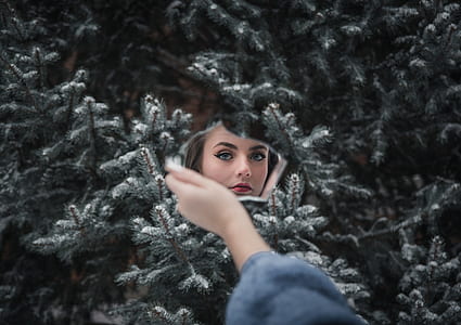 woman in blue sweatshirt holding mirror and showing her face standing in front of pine tree with covered of snow thumbnail