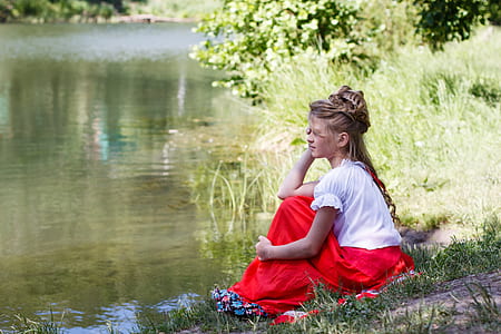 woman wearing white blouse and red maxi skirt sitting on green grass beside body of water thumbnail
