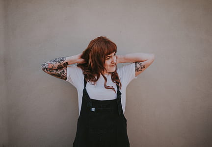 brown haired woman in black dungaree and white shirt standing near wall thumbnail