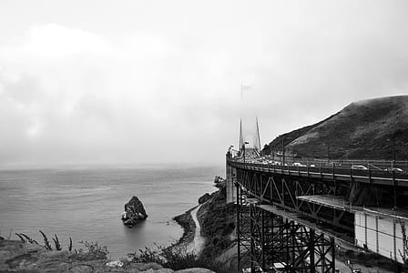 grayscale photography of bridge beside mountain near calm body of sea at daytime thumbnail