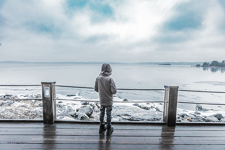 person in gray hoodie standing beside of fence in front of body of water during daytime thumbnail