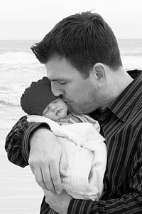 grayscale photo of man kissing baby wrapped in towel on seashore thumbnail