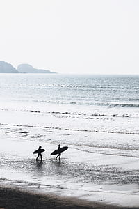 two person holding surfboards walking beside body of water thumbnail
