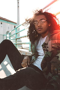 man wearing white crew-neck shirt and camouflage jacket sitting on concrete floor thumbnail