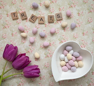 Treats scrabble letters on white, green, and pink floral surface beside chocolate eggs and heart-shaped white ceramic bowl thumbnail