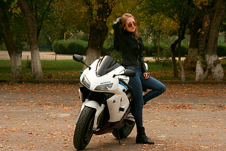 woman wearing blue fitted jeans and black zip-up jacket standing beside white Honda sportsbike thumbnail
