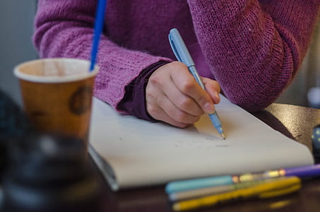 selective focus photography of person holding pen writing on paper on table near coffee cup thumbnail