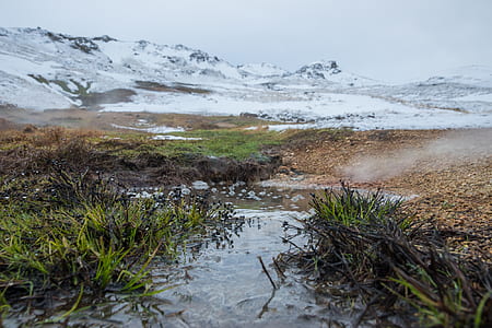 body of water and green grasses at the foot of snow covered mountain during daytime thumbnail