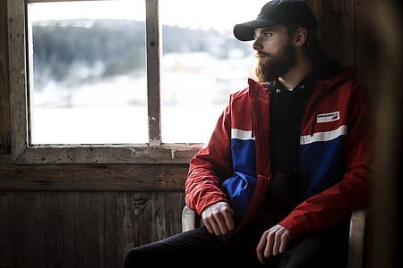 Photo of Man Wearing Black Hat and Blue-and-red Jacket Near Window thumbnail