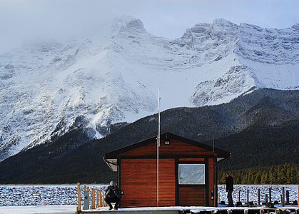 two person on wooden dock with house near body of water and snow covered mountain thumbnail