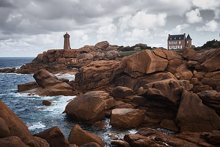 house and tower on the hill surrounded by beige boulders by the sea under white and blue cloudy daytime sky thumbnail