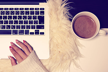 person wearing white sweater with red and white manicure on MacBook Pro beside white ceramic mug with brown liquid thumbnail