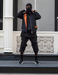 person wearing black balaclava and black and orange jacket standing near the white column thumbnail