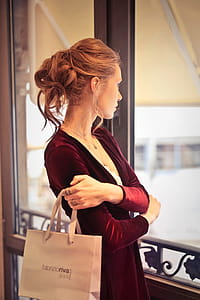 A young blond woman with elegant hair bun wearing a satin dress holding shopping bag in her hand looks out of the windows thumbnail