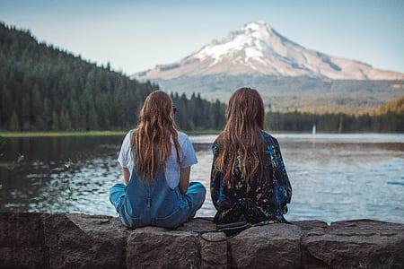 two woman sitting on rock near body of water thumbnail