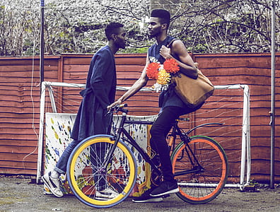 man on bicycle carrying bag with flowers beside man standing near goalie net thumbnail