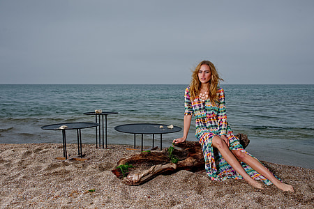 woman sitting on brown driftwood wearing long-sleeved dress on seashore during daytime thumbnail