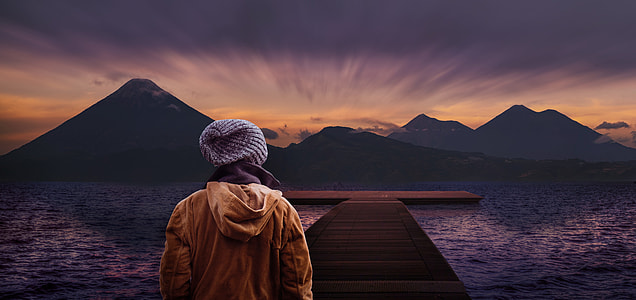 man in gray knit cap standing on brown wooden pathway between body of water during sunet thumbnail