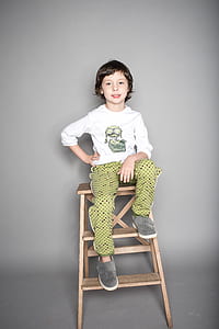 boy wearing white shirt and green bottoms sitting on brown A-frame ladder smiling thumbnail